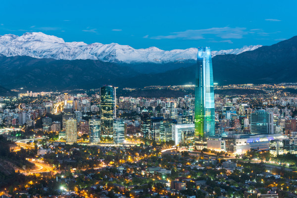 Foto aérea da cidade de Santiago, capital do Chile, repleta de edifícios e com uma cadeia de montanhas ao fundo.
